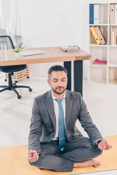 Schöner Geschäftsmann meditiert im Büro in Lotus-Pose auf Fitnessmatte — Stockfoto