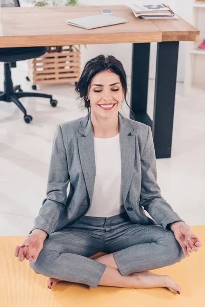 Beautiful smiling businesswoman in suit meditating in Lotus Pose in office — Stock Photo