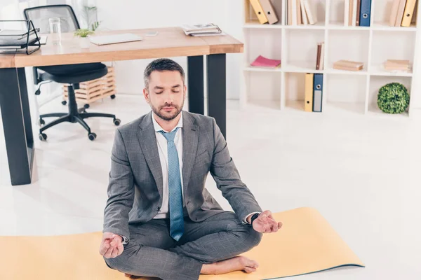 Homem de negócios bonito meditando em Lotus Pose no tapete de fitness no escritório — Fotografia de Stock