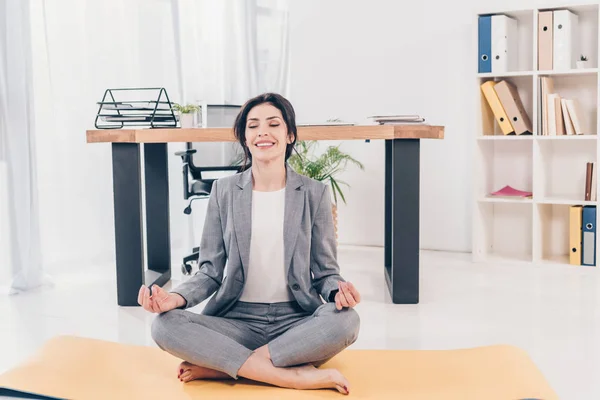 Schöne lächelnde Geschäftsfrau im Anzug sitzt auf Fitnessmatte und meditiert im Büro — Stockfoto