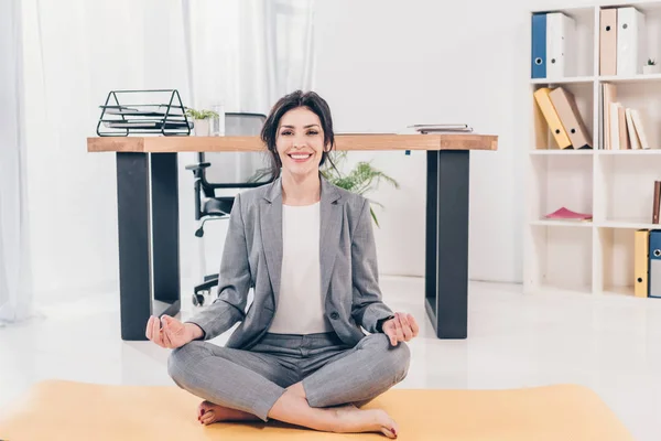 Beautiful smiling businesswoman in suit sitting on fitness mat, looking at camera and meditating in office — Stock Photo