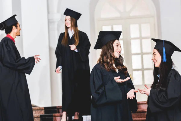 Вибірковий фокус веселих дівчат у випускних сукнях розмовляють, стоячи в університеті — стокове фото