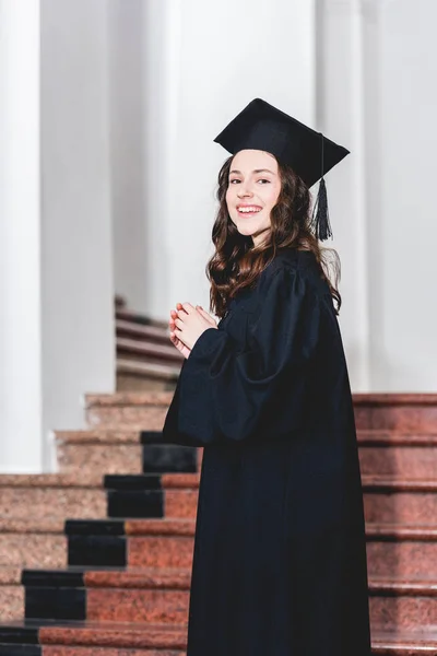 Cheerful young woman in graduation cap smiling while standing in university — Stock Photo