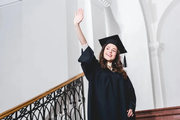 Веселая молодая женщина машет рукой и улыбается в выпускном колпаке — стоковое фото