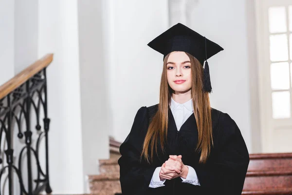 Jovem mulher atraente em boné de graduação olhando para a câmera e de pé com as mãos apertadas — Fotografia de Stock