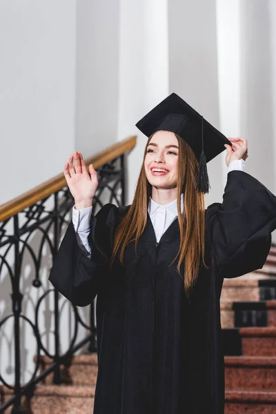Atractiva mujer joven saludando de la mano y sonriendo en la gorra de graduación - foto de stock
