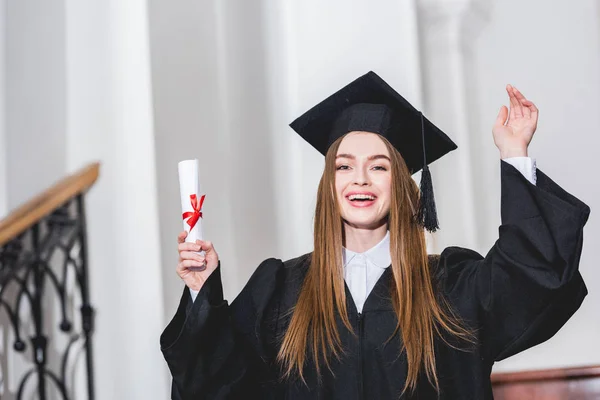 Alegre joven mujer sosteniendo diploma y saludando la mano en la universidad - foto de stock