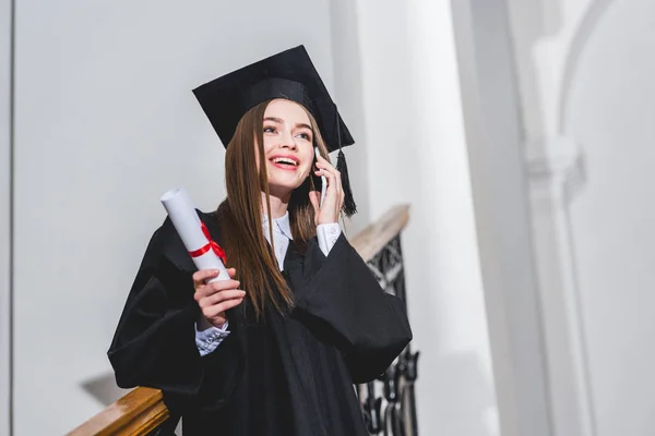 Tiefansicht einer fröhlichen jungen Frau mit Diplom, während sie mit dem Smartphone spricht — Stockfoto