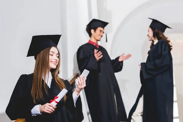 Enfoque selectivo de chica atractiva mirando el teléfono inteligente mientras sostiene el diploma cerca de los estudiantes — Stock Photo