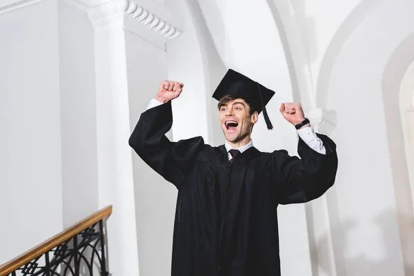Glücklicher junger Mann mit Diplommütze lächelt und gestikuliert in der Universität — Stockfoto