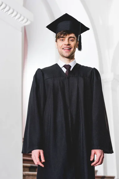 Feliz joven en gorra de graduación sonriendo y mirando a la cámara en la universidad - foto de stock