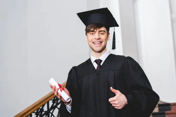 Happy student in graduation gown smiling while holding diploma and showing thumb up — Stock Photo