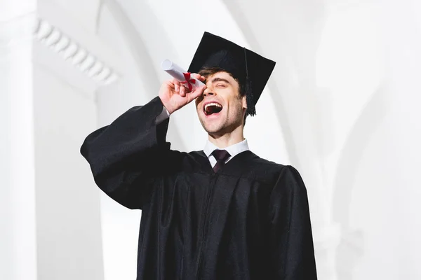 Vista de ángulo bajo de estudiante feliz en vestido de graduación sonriendo mientras sostiene diploma cerca de ojo - foto de stock