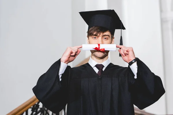 Studentin im Abschlusskleid bedeckt Gesicht mit Diplom — Stockfoto