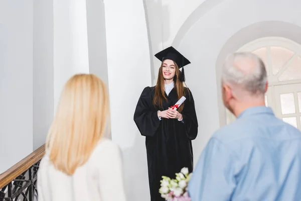 Fuoco selettivo di ragazza allegra in berretto di consegna delle lauree guardando i genitori e il possesso di diploma — Foto stock