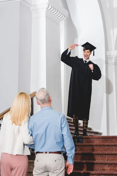 Rückansicht der Eltern, die ihren glücklichen Sohn in der Abschlussmütze gestikulierend mit dem Diplom betrachten — Stockfoto