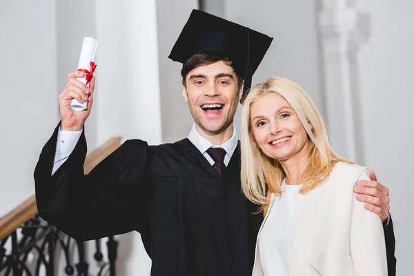 Cheerful son in graduation cap holding diploma while standing with mother — Stock Photo
