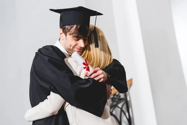 Cheerful son in graduation cap holding diploma while hugging blonde mother — Stock Photo