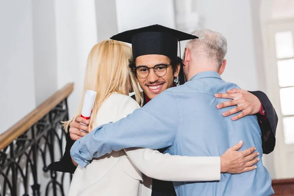 Vista trasera de los padres maduros abrazando al hijo feliz en gafas y gorra de graduación - foto de stock