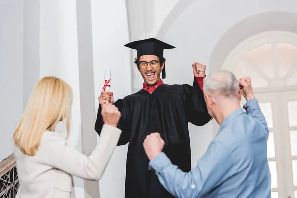 Вид ззаду батьків жестикулює біля щасливого сина у випускній шапці, що тримає диплом — стокове фото