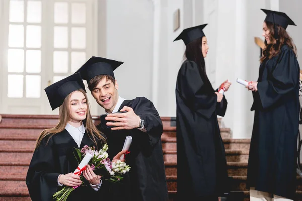 Selektiver Fokus eines gut aussehenden Mannes und eines hübschen Mädchens mit Blumen beim Selfie in der Nähe von Studenten in Abschlusskleidern — Stockfoto