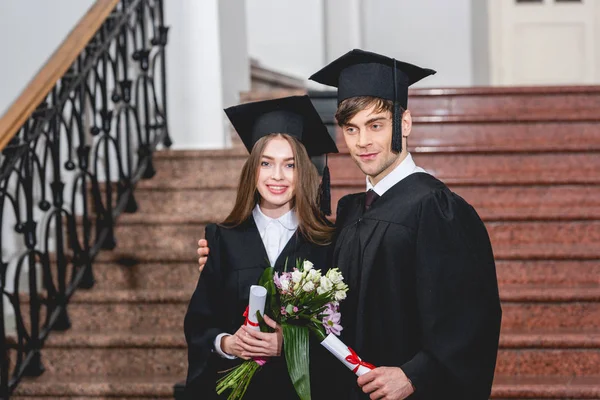 Handsome man and attractive girl with flowers smiling while holding diplomas — Stock Photo