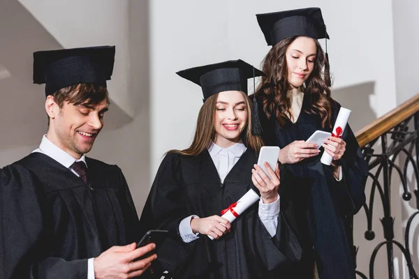 Cheerful students in graduation gowns using smartphones while holding diplomas — Stock Photo