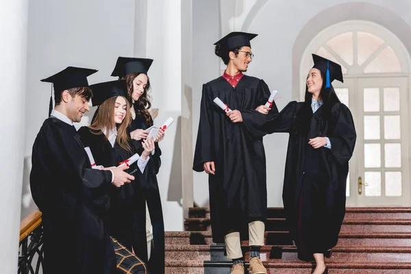 Allegro giovane donna e uomo in possesso di diplomi vicino agli studenti in berretti di laurea utilizzando smartphone — Foto stock