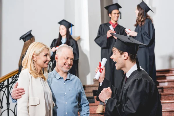 Selective focus of cheerful parents looking at happy son in graduation cap gesturing while holding diploma near students — Stock Photo