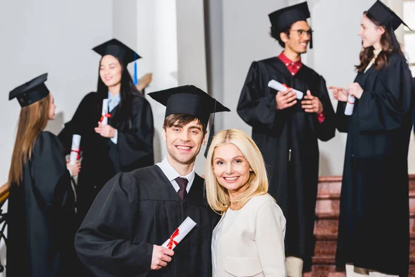 Selective focus of mother and son in graduation cap hugging near students — Stock Photo