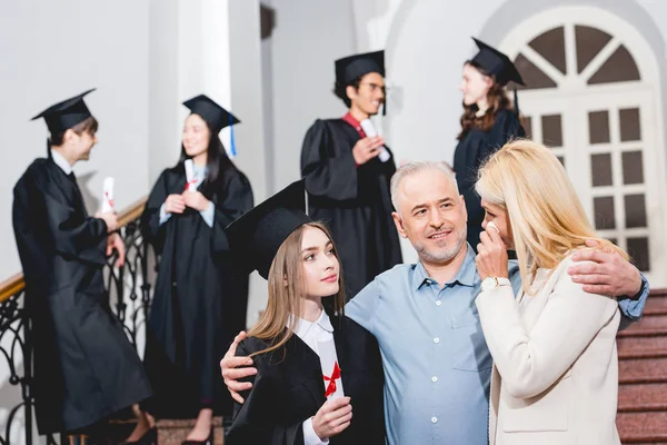 Enfoque selectivo de chica atractiva en la gorra de graduación mirando a la madre llorando mientras se abraza con el padre cerca de los estudiantes - foto de stock