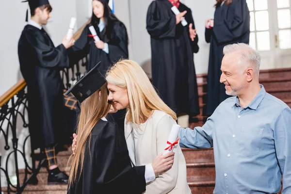 Selective focus of girl in graduation cap hugging with mother near father — Stock Photo