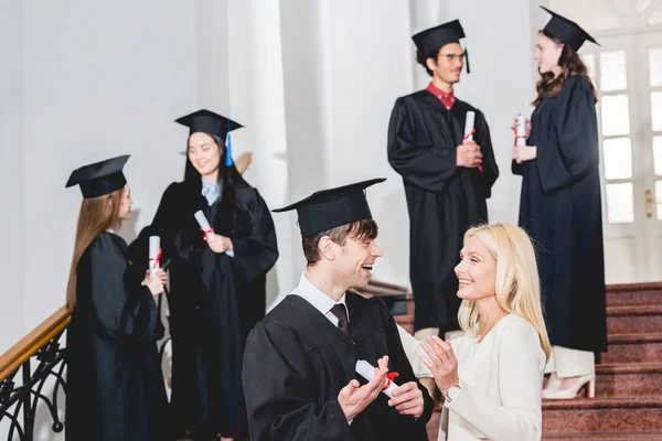 Selective focus of happy son in graduation cap looking at mother near students — Stock Photo