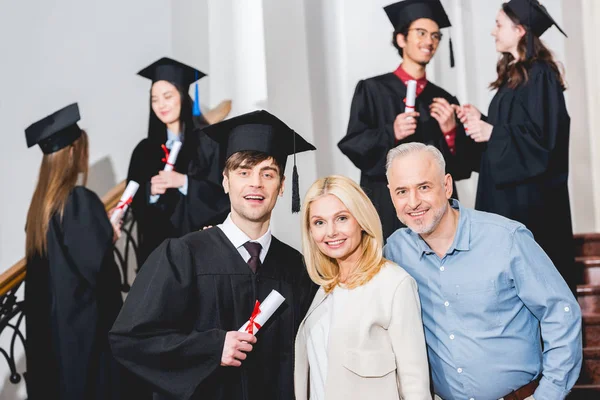 Избирательный фокус счастливого сына в выпускной шапке, стоящего рядом с веселыми родителями — стоковое фото