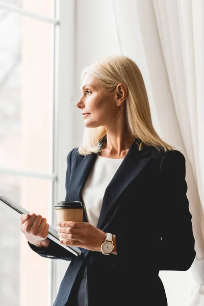 Attraktive blonde Lehrerin steht am Fenster und hält Pappbecher und Ordner in der Hand — Stockfoto