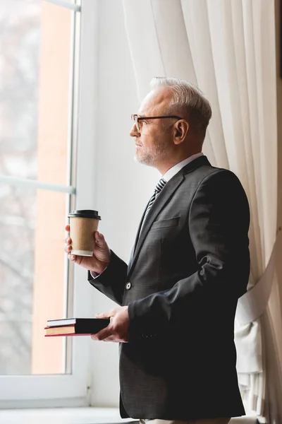 Pensive mature teacher in glasses holding book and paper cup while looking at window — Stock Photo