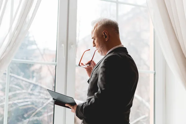 Professeur mature en tenue formelle portant des lunettes et un livre de lecture près de la fenêtre — Photo de stock