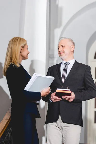 Low angle view of happy mature teacher holding books and looking at attractive colleague with folder in hands — Stock Photo