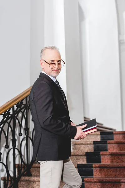 Schöner reifer Lehrer mit Brille, der Bücher hält und in die Kamera schaut — Stockfoto