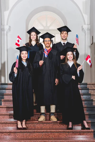 Estudiantes alegres en vestidos de graduación con banderas de diferentes países - foto de stock