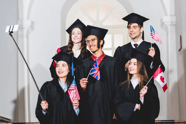 Estudiantes alegres en vestidos de graduación con banderas de diferentes países y tomar selfie en el teléfono inteligente - foto de stock