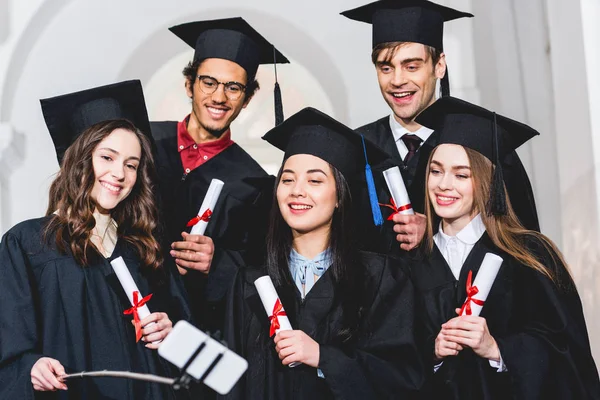 Cheerful group of students in graduation gowns holding diplomas while talking selfie — Stock Photo