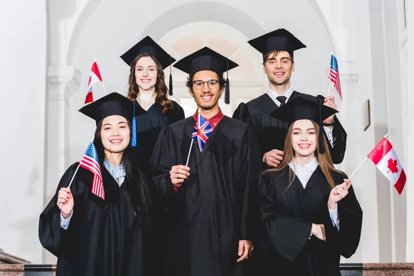 Happy students in graduation gowns holding flags of different countries — Stock Photo
