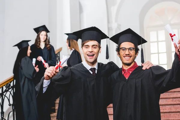 Fuoco selettivo di uomini allegri in berretti di consegna delle lauree che tengono diplomi vicino a studenti — Foto stock