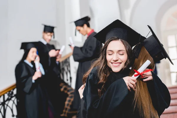 Enfoque selectivo de chica alegre sosteniendo diploma y abrazando cerca de los estudiantes - foto de stock