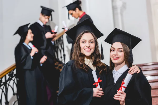 Enfoque selectivo de hermosas chicas en gorras de graduación sonriendo mientras sostiene diplomas cerca de los estudiantes - foto de stock