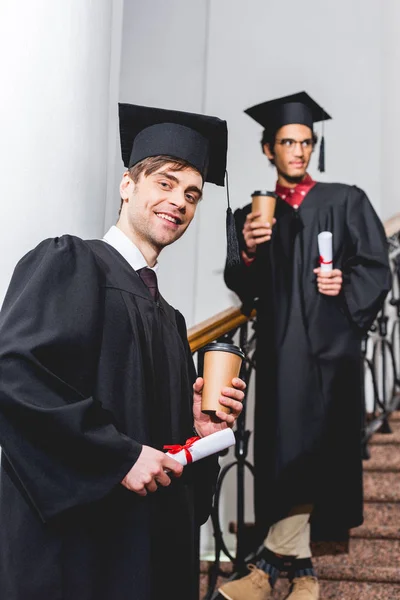 Messa a fuoco selettiva di uomo allegro in berretto laurea in possesso di diploma e tazza di carta vicino studente — Foto stock