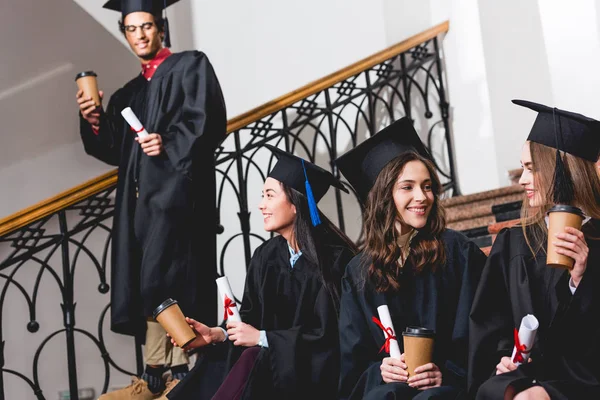 Estudiantes alegres y atractivos sosteniendo vasos de papel y diplomas cerca del estudiante - foto de stock