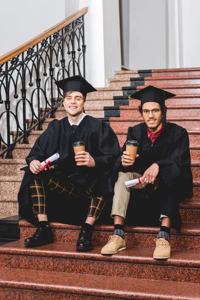 Fröhliche Studenten im Abschlussgewand mit Urkunden und Pappbechern auf der Treppe — Stockfoto