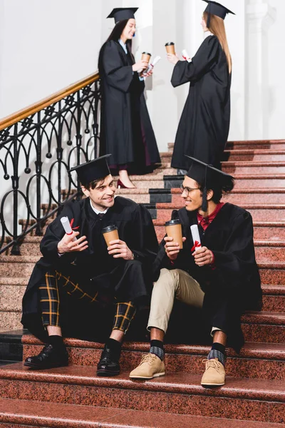 Messa a fuoco selettiva di studenti allegri in abiti di laurea in possesso di diplomi e bicchieri di carta mentre seduto sulle scale — Foto stock